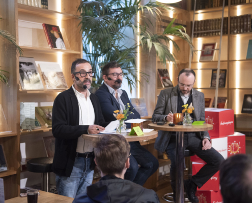 en panelsamtale med tre menn som sitter ved høye bord i et bibliotek