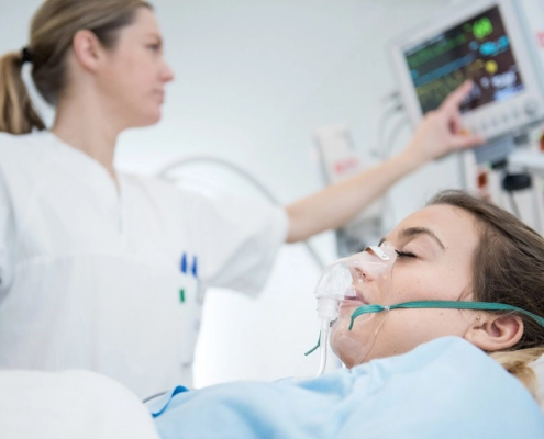 En pasient som ligger i sykeseng med pustemaske. I bakgrunnen ser vi en sykepleier som sjekker en hjertemonitor.