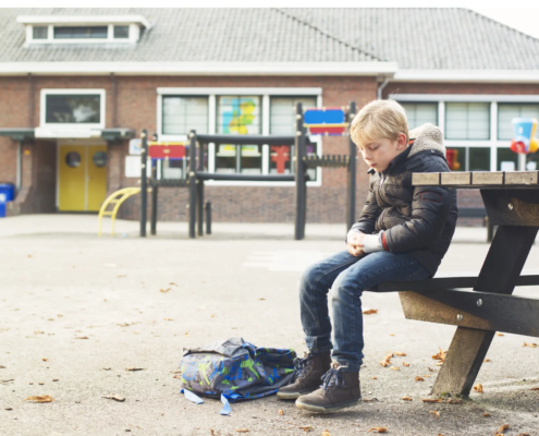 Gutt som sitter på en benk i en skolegård. Ser ensom ut og sekker ligger ved føttene hans.
